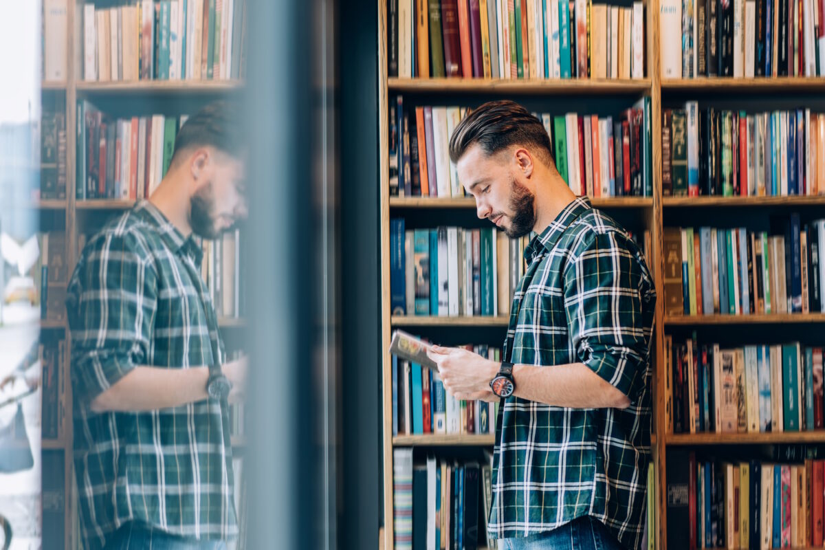 Side view of Caucasian male student in casual clothing reading best seller novel in public bookstore