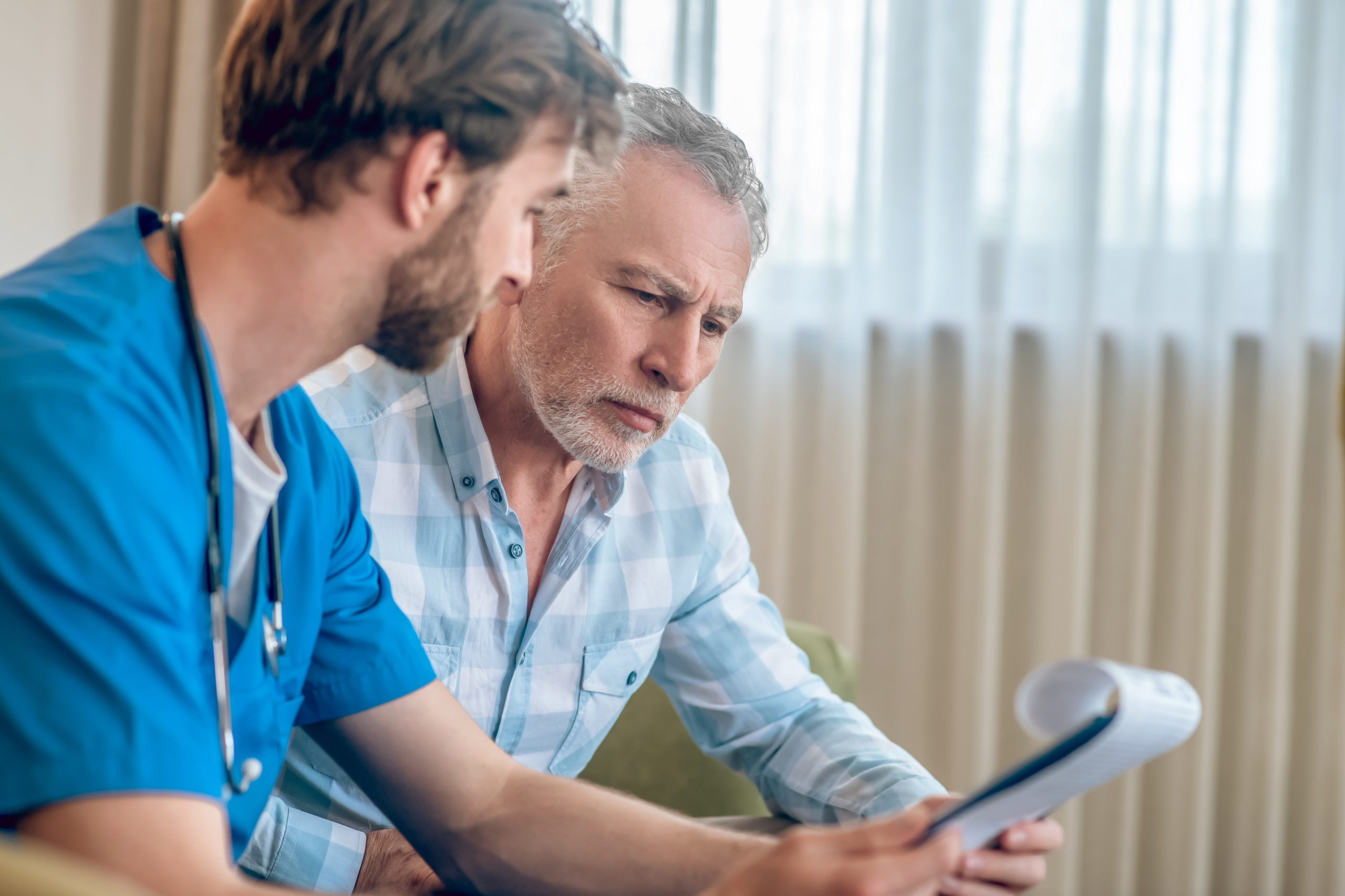 focused-serious-gray-haired-man-plaid-shirt-looking-clipboard-doctor-hands-stroke-scaled