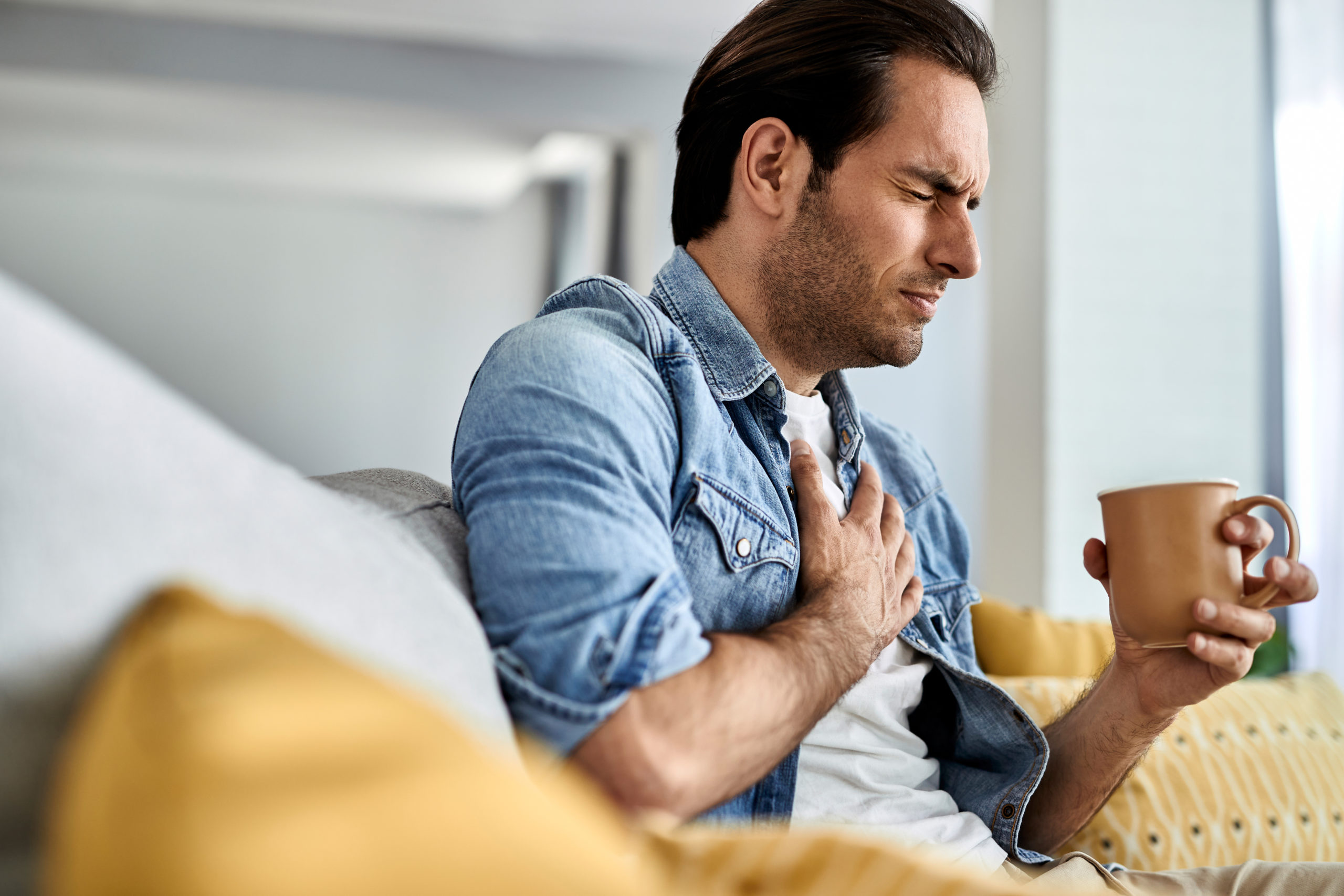 young-man-feeling-sick-holding-his-chest-pain-while-drinking-tea-living-room-beat-heartburn-scaled