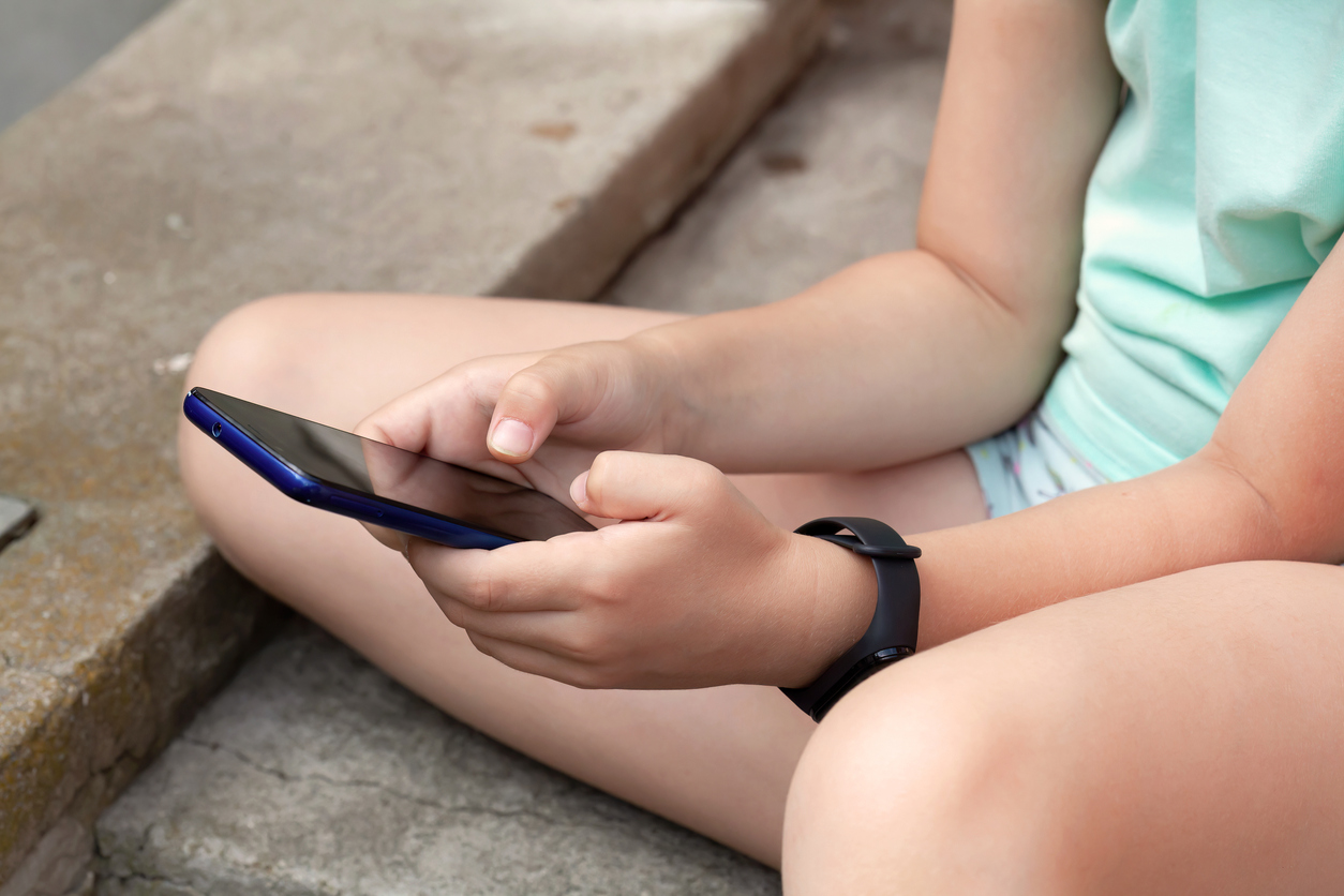 Anonymous child sitting outdoors using a smartphone outdoors, hands closeup detail shot, face obscured, girl typing and scrolling social media on mobile device, wearing smart band fitness tracker