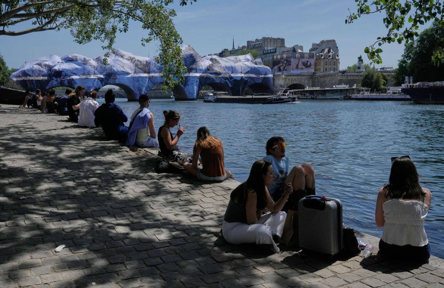 Πολίτες αναζητούν δροσιά στις όχθες του Σηκουάνα στο Παρίσι, ενώ στο βάθος διακρίνεται έργο του street artist JR στη γέφυρα Pont Neuf. Φωτογραφία: AP Photo/Michel Euler