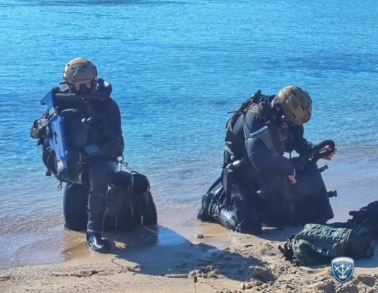Δύο στρατιωτικοί δύτες με εξοπλισμό κατάδυσης και κράνη γονατιστοί στην ακτή