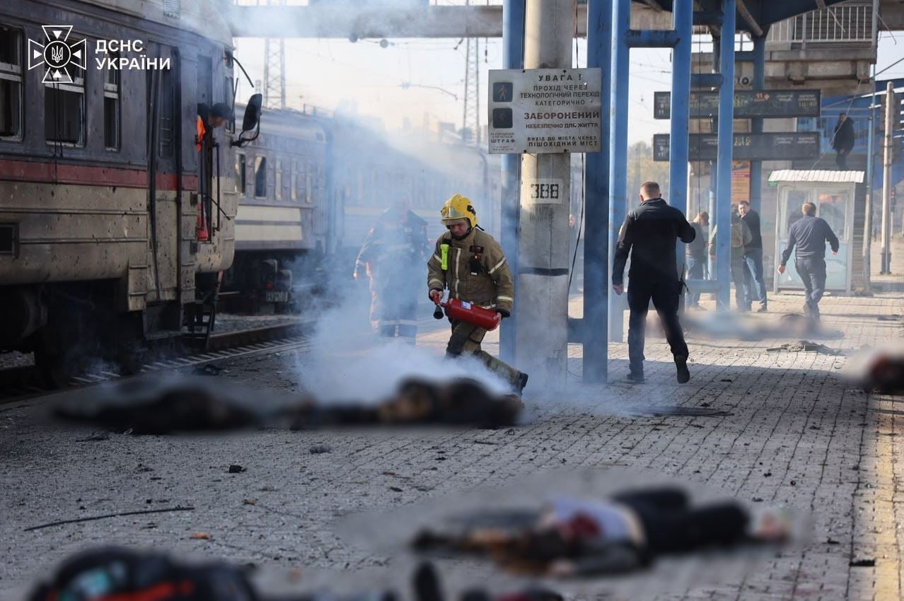 Καπνοί καλύπτουν την αποβάθρα του σιδηροδρομικού σταθμού στο Ντνίπρο, ενώ πυροσβέστες επιχειρούν δίπλα σε επιβατική αμαξοστοιχία και στο έδαφος διακρίνονται οι θολωμένες σοροί θυμάτων της ρωσικής επίθεσης. (Φωτογραφία: DSNS Ουκρανίας)
